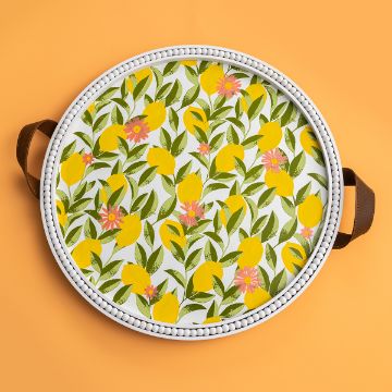 Show details for Citrus Trees A round serving tray with white beaded edges and brown leather handles is decorated with a colorful pattern of yellow lemons, green leaves, and pink flowers on a white background. The tray is set against a bright orange surface featuring the Chalk Couture transfer.