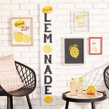 Show details for Fresh Lemonade A white brick wall displays a vertical sign reading "Fresh LEMONADE" alongside several lemon-themed framed prints. Two lemonade glasses with straws, and lemon slices sit on a round tray on a black chair. An orange-patterned pillow rests on another chair.