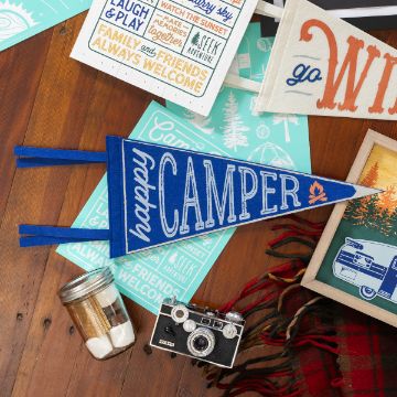 Show details for Felt Pennant Flag (Blue, 17 x 7 1/5) A wooden surface displays camping-themed items, including a blue pennant with "happy CAMPER" text, a vintage camera, a mason jar with small white objects, and assorted signs with messages like "Family and Friends Always Welcome." A plaid blanket is partially visible.