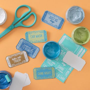Show details for Coupon An orange surface with various colorful paper tickets, open paint containers with blue, white, and green paints, and a pair of teal scissors. Tickets include phrases like "Just Us," "Car Wash," and "Game Night." Chalk Couture transfer is also present.