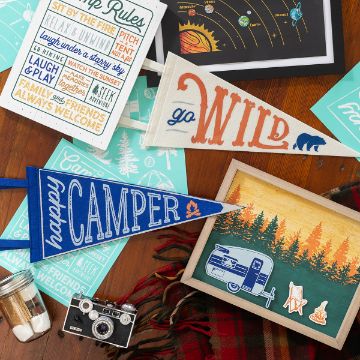 Show details for Go Wild A colorful flat lay features vintage camping-themed items, including pennant flags that say, "Go Wild" and "Happy Camper," a framed artwork of a camper in a forest, a retro camera, and various Chalk Couture transfers with outdoor adventure quotes.