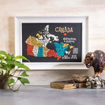 Show details for Map of Canada A framed chalkboard map of Canada highlights its provinces and territories in various colors. The transfer by Chalk Couture is displayed on a table beside a green plant, two old books, and a buffalo figurine. The background wall is light-toned.