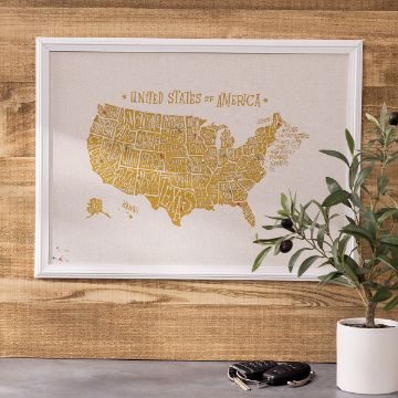 Show details for Map of United States A framed map of the United States, featuring each state's name in yellow typographic lettering, created using the "transfer" by Chalk Couture, hangs on a wooden wall. A small potted plant and a set of keys with a keychain sit on a white table below the map.