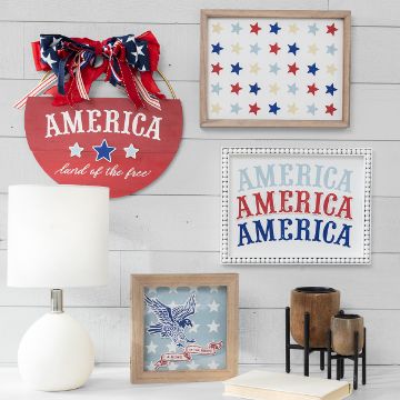 Show details for Land of The Free A white wall is decorated with July 4th-themed items: a "Land of the Free" sign topped with red and blue bows, star patterns created using a Chalk Couture transfer, and "America" repeated. Below are a white lamp, framed eagle art, and a set of three wooden vases on a black stand.