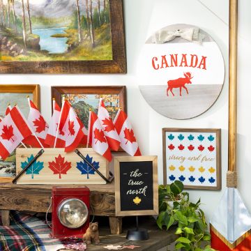 Show details for The True North A shelf featuring Canadian-themed decorations including small Canadian flags, framed prints with maple leaves, a circular sign with a moose and the word "Canada," a framed quote "the true north," a framed needlepoint "strong and free," and a hanging paddle designed using the Chalk Couture transfer.