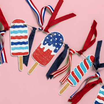 Show details for Popsicles Chalkable Shapes (3 Pieces) A garland featuring popsicle-shaped decorations in red, white, and blue patriotic designs hangs against a pink background. The garland is also adorned with red, white, and blue ribbons.