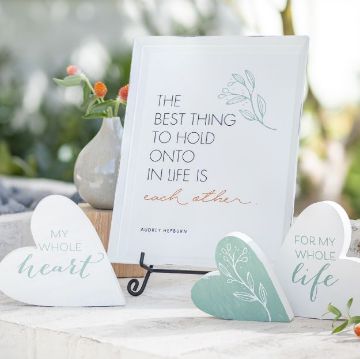 Show details for My Whole Heart A framed Chalk Couture transfer, "The best thing to hold onto in life is each other," stands on a metal stand. Nearby, two heart-shaped signs read "For my whole heart" and "For my whole life". They sit on a white surface with potted flowers in the background.