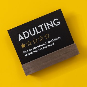 Show details for Reusable Black Board & Base (5" x 7") A black sign with white text on a wooden stand against a yellow background. The sign reads "ADULTING" in large letters, followed by 1 out of 5 stars filled in, and the text beneath says, "Not as advertised. Definitely would not recommend.