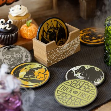 Show details for Coaster Set (Black, 7 Pieces) A collection of Halloween-themed coasters featuring various spooky illustrations is displayed on a table. The coasters include designs such as a bat, a witch, and a "Trick or Treat" message. Nearby are decorated cupcakes and a green glass container.