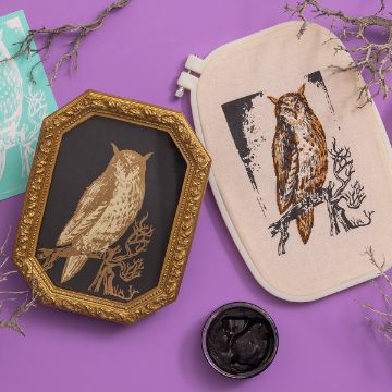 Show details for Wise Owl A decorated workspace with a framed gold and black owl print on the left and an embroidered owl in a hoop on the right. The background is purple with twiggy branches scattered around, and there is a stencil and black paint jar also visible.