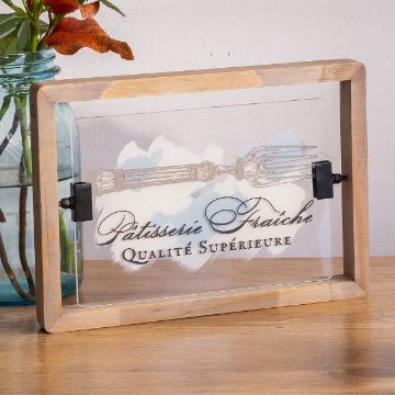 Show details for Bistro Café A wooden frame holds a glass panel featuring the text "Pâtisserie Fraîche Qualité Supérieure" and an illustration of a fork. The backdrop is a muted cloud-like pattern. The frame rests on a wooden surface beside a glass jar with orange flowers.