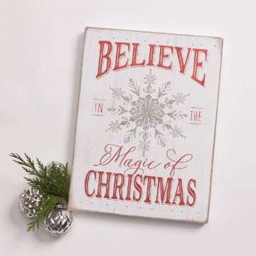 Show details for Believe In The Magic A wooden sign with festive lettering that reads "Believe in the Magic of Christmas" in red and silver, adorned with a silver snowflake design in the center. The sign is complemented by sprigs of greenery and two silver ornaments beside it.
