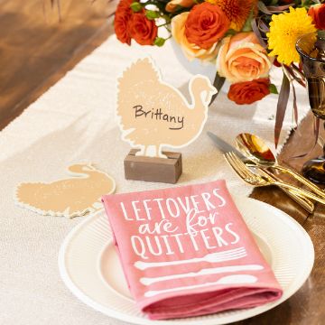 Show details for Feast Mode A table setting with a white plate and a pink napkin with the phrase "LEFTOVERS ARE FOR QUITTERS" in white letters. A decorative turkey card with "Brittany" written on it stands at the top of the plate. The table is adorned with an orange and yellow flower arrangement.
