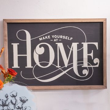 Show details for At Home A framed sign with a black background and elegant white lettering that says, "Make Yourself At Home." The decorative text features curves and flourishes. In the bottom left corner, there are some red and white flowers partially visible.