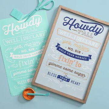 Show details for Hold Your Horses A framed art piece and printed stencil featuring Southern-themed phrases like "Howdy y'all", "well I declare", "yes, ma'am", "hold your horses", "fixin' to", "gimme some sugar", and "bless your heart". Beside them are scissors and an orange squeegee on a turquoise surface.