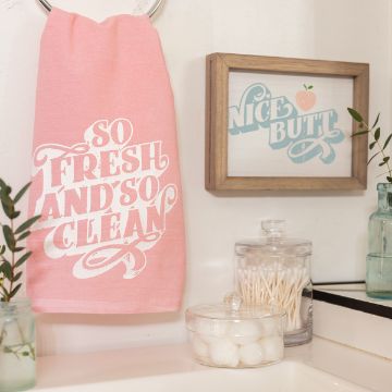 Show details for So Fresh A bathroom scene with a pink towel on a ring that says "So Fresh and So Clean" in white text. Beside it is a framed sign that says "Nice Butt" with an image of a peach. There are jars of cotton balls and cotton swabs on the counter, plus some green plants.