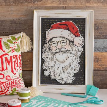 Show details for Believe Santa A framed picture of Santa Claus with a jolly face and white beard, wearing glasses and a red hat. The frame is set against a rustic wooden background. To the left is a festive pillow with the text "Christmas Tidings," and craft supplies are scattered around.