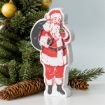 A wooden cutout decoration of Santa Claus holding a gift sack is displayed in front of a Christmas tree with golden ornaments and pinecones. Santa is depicted in his traditional red suit and hat, with white trim and a black belt and boots.