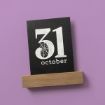 A black card displaying "31 October" with a white spider web design within the number "3" is held upright by a wooden stand on a purple background.