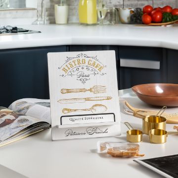 Show details for Cookbook Stand A kitchen with a cookbook stand displaying a sign that reads "Bistro Cafe Paris" with illustrations of cutlery. The counter has measuring cups, a bowl, a kettle, and some vegetables in the background, including tomatoes and chili peppers.