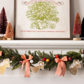 Show details for Christmas Angel A festive mantle decorated with a lush green garland adorned with red, gold, and silver ornaments and peach-colored bows. Paper angel ornaments hang from the garland. Above the mantle, a framed holiday-themed artwork is flanked by small, colorful bottle brush trees.