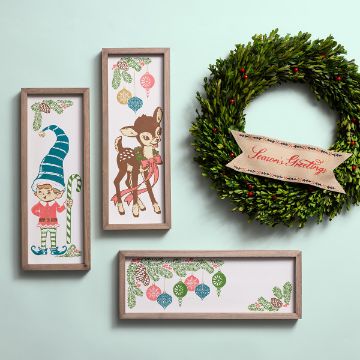 Show details for Pine & Ornaments A holiday-themed display features three framed artworks: an elf holding a candy cane, a fawn with a scarf, and hanging ornaments. A green wreath with red berries and a "Season's Greetings" ribbon is mounted beside them, all set against a pale green wall.