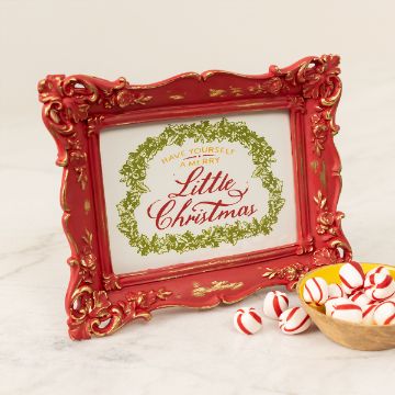 Show details for Little Christmas A red ornate picture frame with gold detailing sits on a white surface. Inside the frame, text reads "Have Yourself a Merry Little Christmas" surrounded by a green wreath. Nearby, a wooden bowl contains red and white striped peppermint candies.