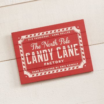 Show details for Candy Cane Factory A red sign with a vintage design that reads, "The North Pole Candy Cane Factory. Old fashioned, hand-rolled. Hand delivered by Kris Kringle." The text is in a mix of white and decorative fonts, framed by a candy cane border. The sign is hanging on a white wall.