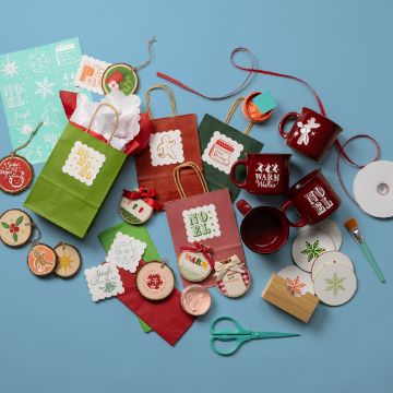 Show details for From Santa With Love A collection of Christmas-themed craft supplies and gift items on a blue background. Includes gift bags, mugs, coasters, stickers, ornaments, ribbon, and crafting tools like scissors and a paintbrush. Predominantly red, green, and festive designs.