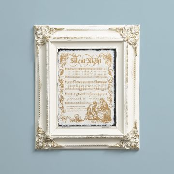 Show details for Vintage Silent Night A framed sheet music piece for "Silent Night" is centered on a light blue wall. The ornate white frame has intricate floral details at the corners. The sheet music is decorated with golden patterns, and features an illustration of two figures at the bottom.