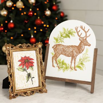 Show details for Vintage Deer A festive scene features a framed poinsettia illustration and a wooden stand displaying a circular plaque with a reindeer and green leaves. Behind them, a Christmas tree adorned with red and gold baubles is visible. The background is a light-colored wall.
