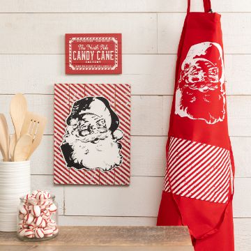 Show details for Santa Claus Decorative kitchen set featuring a red apron with a white Santa Claus print, a matching striped wall sign with Santa Claus, a smaller "Candy Cane Lane" sign, wooden cooking utensils in a white holder, and a glass jar filled with peppermint candies.
