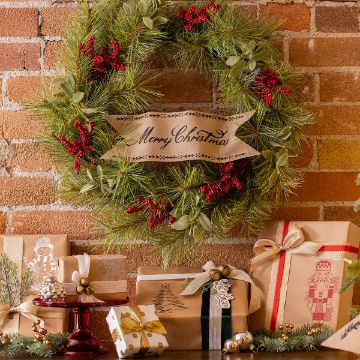 Show details for Sleigh Bells Ring A festive Christmas wreath with red berries and a "Merry Christmas" ribbon is hung on a brick wall. Below it, a variety of wrapped gifts with ribbons and festive decorations are arranged on a table, creating a cheerful holiday scene.
