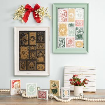 Show details for Holiday Stamps A festive display featuring framed Christmas stamps hung on a wall. One frame has black and gold stamps, and the other has colorful stamps. A red bow with white berries is above, and a table below holds small holiday cards, red berries, and a string of pearls.