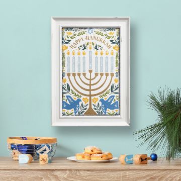 Show details for Hanukkah Menorah A framed "Happy Hanukkah" print featuring a menorah is on a light blue wall. On the wooden table below, there are dreidels, a bowl with blue and white designs, a bunch of evergreen branches, and a plate with doughnuts.