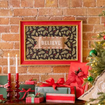 Show details for Shapeable Banner (4¼” x 13½") A cozy Christmas-themed room features a brick wall adorned with a framed sign that says "Believe" surrounded by holly and pine designs. Below are wrapped presents in red and green, red candlesticks with lit white candles, and a partially visible decorated Christmas tree.