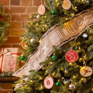 Show details for Shapeable Banner (6¼" x 47”) A Christmas tree decorated with ornaments and a large ribbon that reads "Peace on Earth. Good Will To..." The tree is adorned with green and red ornaments, and a small gift wrapped in festive paper sits on a table nearby. In the background is a brick wall.