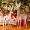Two decorative wooden reindeer figurines with text patterns stand in front of a Christmas tree adorned with lights and ornaments. They are surrounded by wrapped gifts, including one with a gold ribbon and another with a small pinecone on top.