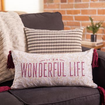 Show details for Lumbar Pillow Cover (White, 12" x 24") A rectangular pillow with red tassels and gold floral patterns, featuring the text "It's a Wonderful Life" in red, sits on a dark gray couch. Behind it is a beige checkered pillow and a beige knitted throw blanket, with a brick wall partially visible in the background.