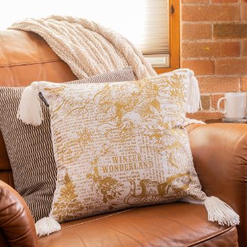 Show details for Square Pillow Cover (2-Pack, White, 17½" x 17½") A tan leather couch adorned with a large pillow. The pillow features intricate gold and white designs, including a "Winter Wonderland" text and Christmas motifs. A white knitted blanket and a striped pillow are on the couch. A mug sits beside, near a brick wall.