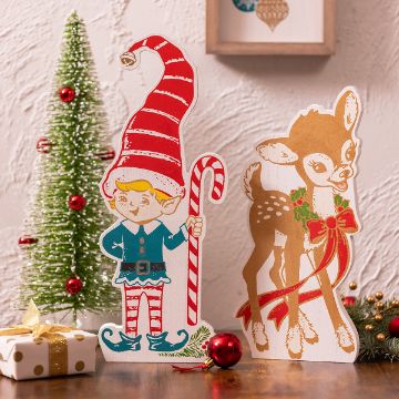 Show details for Elf & Reindeer Chubbies Festive holiday scene featuring a cardboard cutout of an elf with a candy cane and a reindeer adorned with a ribbon. Background includes a decorated Christmas tree, wrapped gift, red ornaments, and framed artwork on the wall.