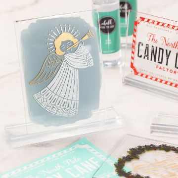 Show details for Table Display Mold (Board & Base Mold (5" x 7") A decorative display with a painted angel blowing a trumpet on a clear stand. Surrounding items include a North Pole Candy Cane Factory sign and other festive accessories, all set on a white background.