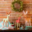 A festive scene featuring three colorful, wooden reindeer figurines in front of a brick wall adorned with a green wreath. Crafts supplies like scissors, tape, and glue are scattered on the table. The reindeer are decorated in green, red, and white holiday patterns.