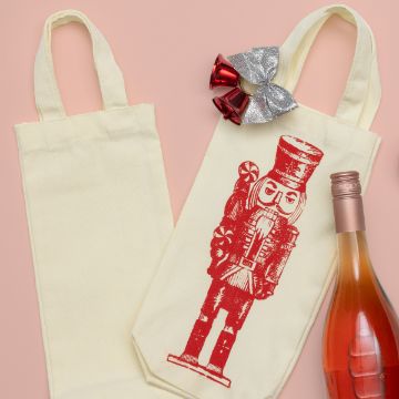 Show details for Wine Bag Two beige wine bags, one with a red nutcracker design, are on a peach background. A bottle of rosé wine is next to them. A silver and red bell decoration is placed on the bag with the nutcracker.