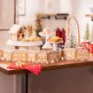 A festive kitchen scene with a gingerbread house and train on a wooden table. Various cookies and treats are arranged on cake stands. A red towel with white polka dots is draped over the table edge. A decorated Christmas tree is visible in the background.