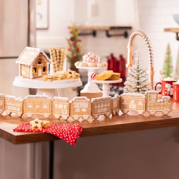 Show details for Camel Paste Single A festive kitchen scene with a gingerbread house and train on a wooden table. Various cookies and treats are arranged on cake stands. A red towel with white polka dots is draped over the table edge. A decorated Christmas tree is visible in the background.