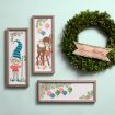 Three vintage holiday-themed framed prints feature an elf, a reindeer, and colorful ornaments. A green wreath with a red "Season's Greetings" banner is displayed on a light green wall.