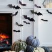 A fireplace with a warm glow is decorated with black bat cutouts on the white wall. Below, a variety of pumpkins in white, green, and purple hues are arranged, adding to the Halloween theme.