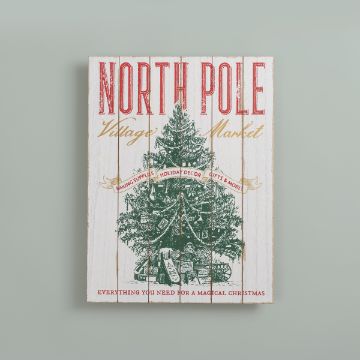 Show details for Shiplap Sign (18" x 24") A festive sign reads "North Pole Village Market" with a vintage-style illustration of a Christmas tree decorated with ornaments, ribbons, and surrounded by presents. The text includes "Holiday Decor" and "Everything you need for a magical Christmas.