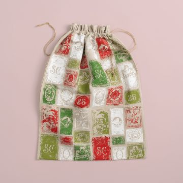 Show details for Drawstring Bag (Tan, 24" x 32") A drawstring bag with a vintage Christmas stamp design in red, green, and white on a soft beige background. Stamps feature Santa, snowflakes, and festive symbols. The bag is set against a light pink surface.